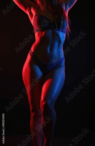 Fit woman in lingerie posing in red and blue neon lights against black backdrop. Her toned body features defined abs and smooth skin illuminated by contrasting colors.