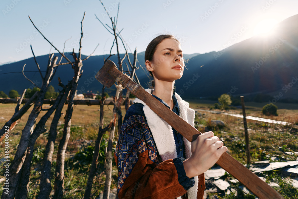Naklejka premium Woman in countryside field holding an axe over shoulder by wooden fence, mountains in background and sunlight. Rustic portrait of a young farmer in rural landscape and nature.