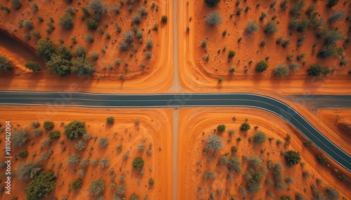 Aerial drone view shows desert roads intersection. Dark paved highway crosses unpaved track in red earth landscape. Sparse green bushes define arid Outback region. Remote, vast expanse, long