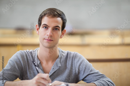 Adult male wearing grey henley sitting at lecture desk, holding pen and writing notes