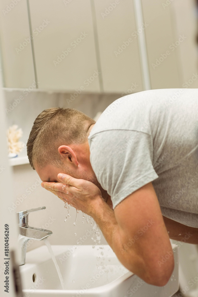 Obraz premium Adult male wearing gray T-shirt washing face at vanity using chrome faucet, water droplets falling