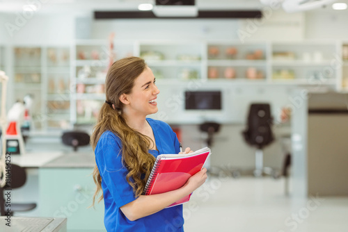Woman in blue scrub top holding red notebook and pink folder in clinical lab, copy space