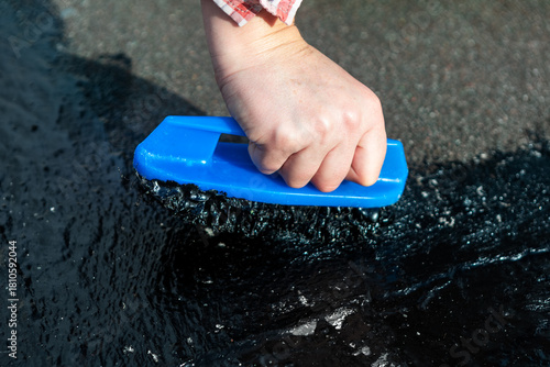 Hand pushes brush through thick black sealant, applying waterproof coating to surface like roof or pavement. Process involves using asphalt or tar compound with mastic brush for sealing and repairing