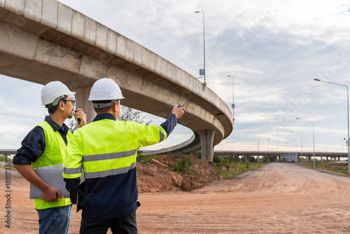 Two construction managers, communicating via radio, inspect a dusty highway site under a curving concrete overpass.