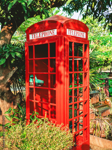 Red vintage London style telephone booth in garden. Traditional British classic telephone booth in park surrounded by lush green and trees. Old fashioned british phone box. Сlassic red telephone box.