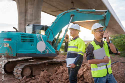 Canvas-taulu Two construction engineers use two-way radios to coordinate work in front of an excavator under a concrete highway overpass