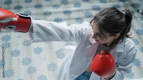 Young woman practicing boxing techniques in a bright gym with patterned flooring, wearing a white coat and red gloves