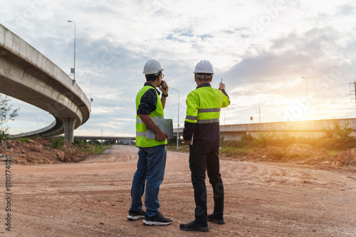 Construction workers discussing project details at sunset, wearing safety vests and helmets, overseeing highway construction site with clouds and asphalt road