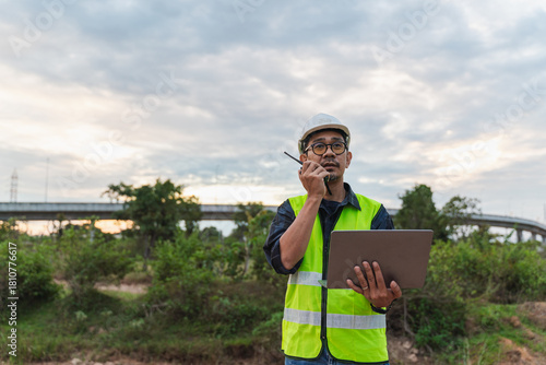 Construction Manager Working Outdoors with Laptop and Walkie-Talkie at Construction Site During Sunset with Bridge in Background