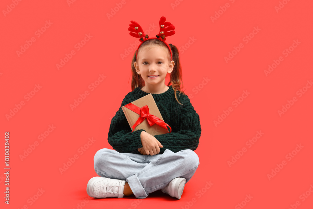Fototapeta premium Cute little girl in Christmas reindeer horns with gift book sitting on red background