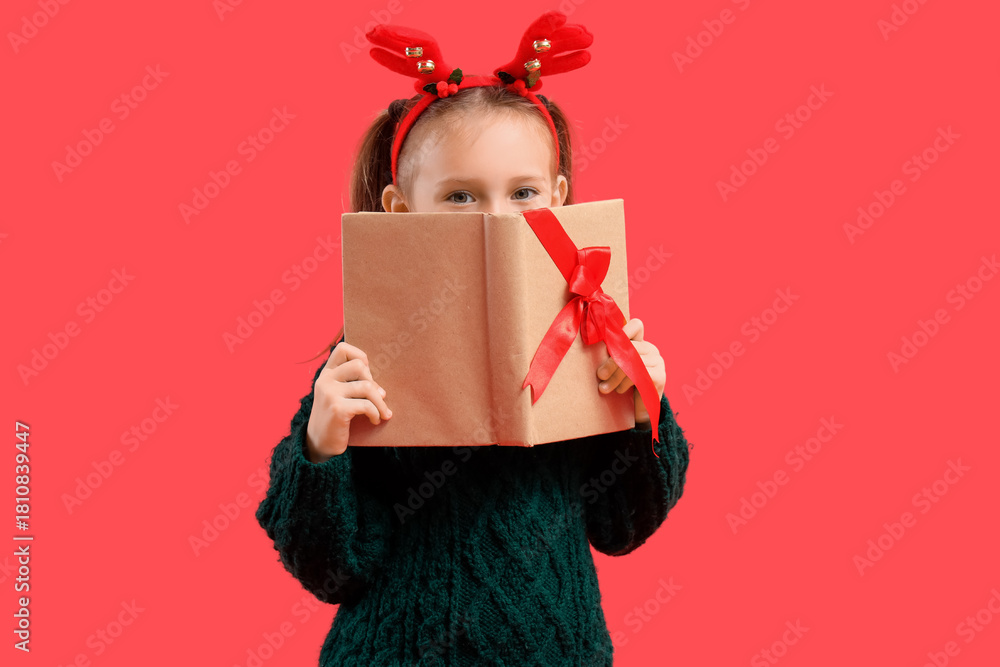Naklejka premium Cute little girl in Christmas reindeer horns with gift book on red background