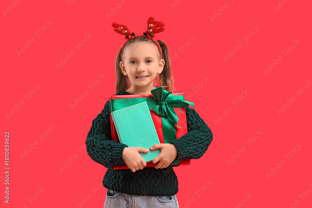 Naklejka premium Cute little girl in reindeer horns with book and Christmas gift on red background