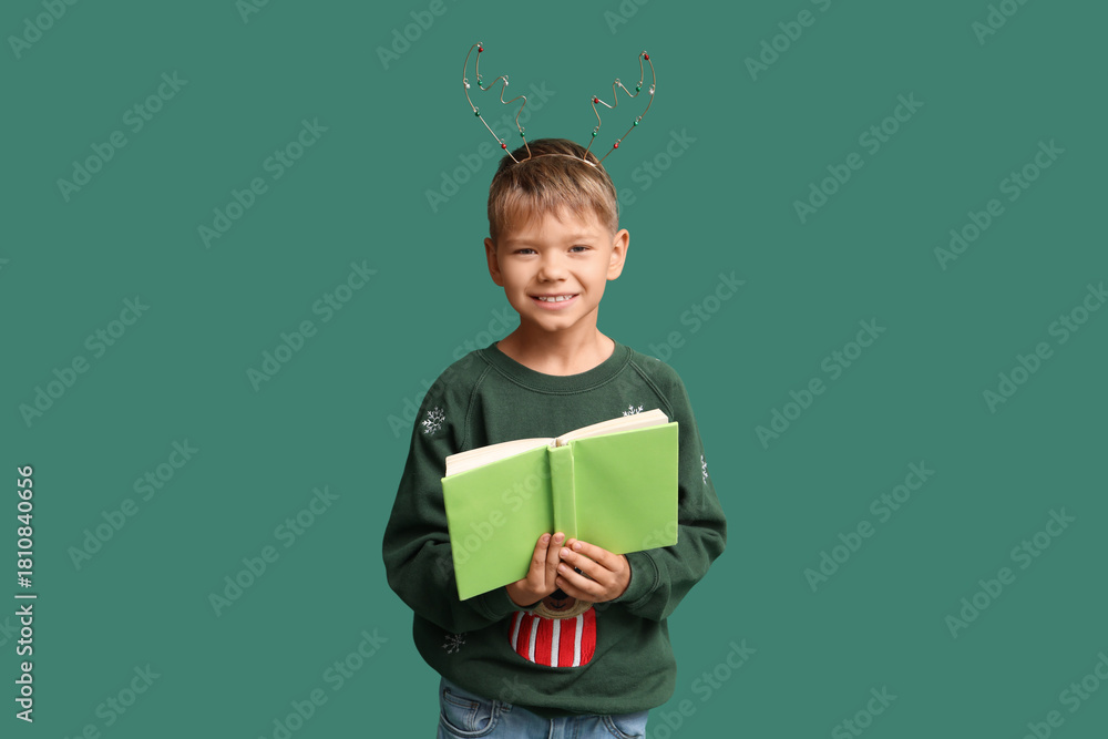 Naklejka premium Cute little boy in Christmas reindeer horns with book on green background