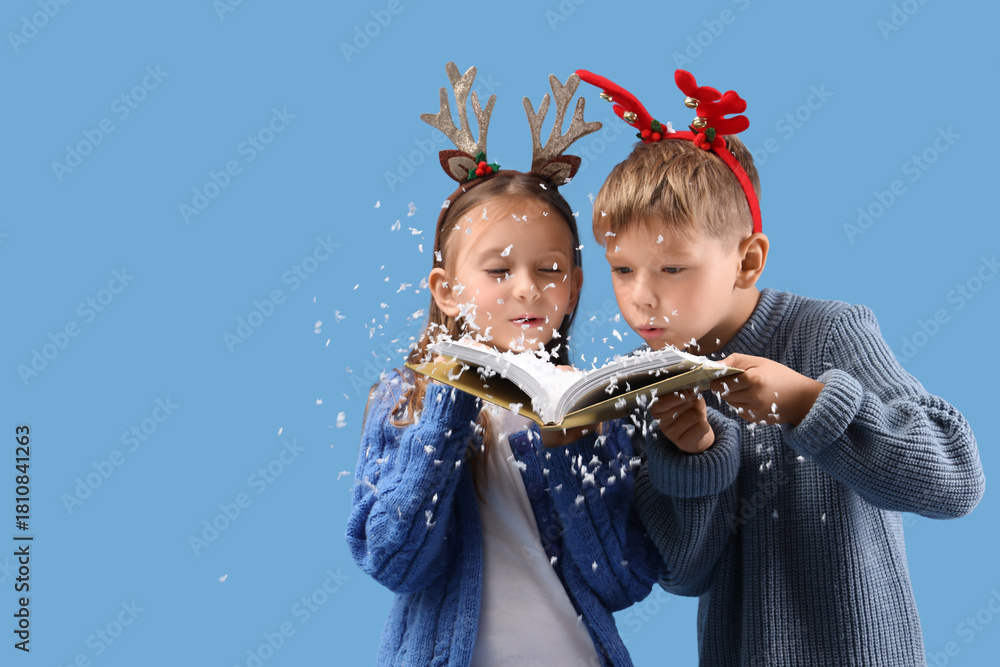Naklejka premium Cute little children in Christmas reindeer horns blowing snow off book on blue background