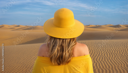 Fototapeta Naklejka Na Ścianę i Meble -  A woman in a yellow hat and shoulderless top overlooks a vast landscape of sand dunes. Calming blue sky.