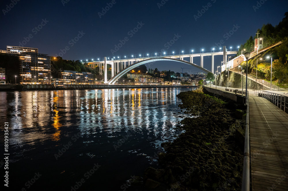 Obraz premium Brightly illuminated Arrabida Bridge and surrounding buildings reflected in calm water of Douro River in Porto, Portugal at night.