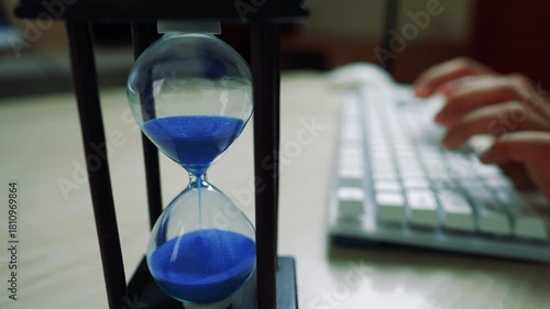 Close-up of an hourglass with blue sand next to a person typing on a keyboard