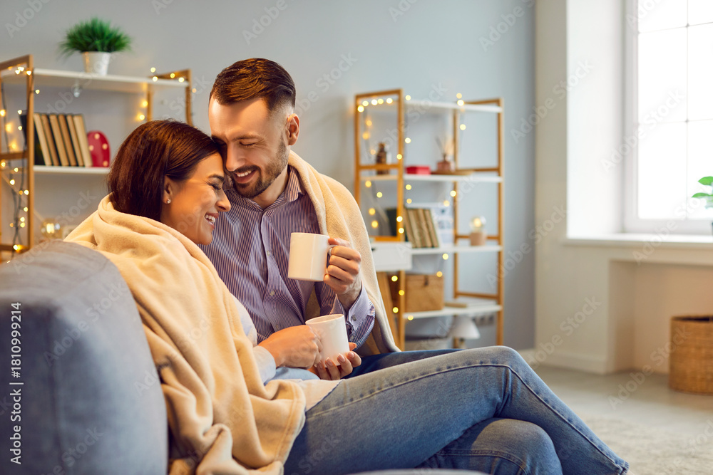 Fototapeta premium Happy cheerful joyful married couple enjoying good relationship, spending winter day off at home, cuddling on comfy cosy sofa in living room with Xmas lights, drinking hot tea, and having fun together