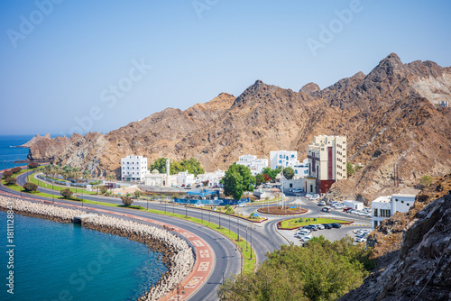 A scenic view of Old Muscat, Oman, featuring a curving coastal road, deep blue water, and dramatic rugged mountains rising behind white traditional buildings