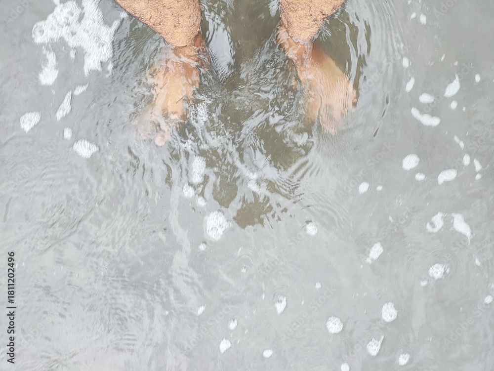 Fototapeta premium A low-angle shot shows bare feet partially submerged in shallow, foamy ocean water that washes over dark sand, creating gentle ripples.