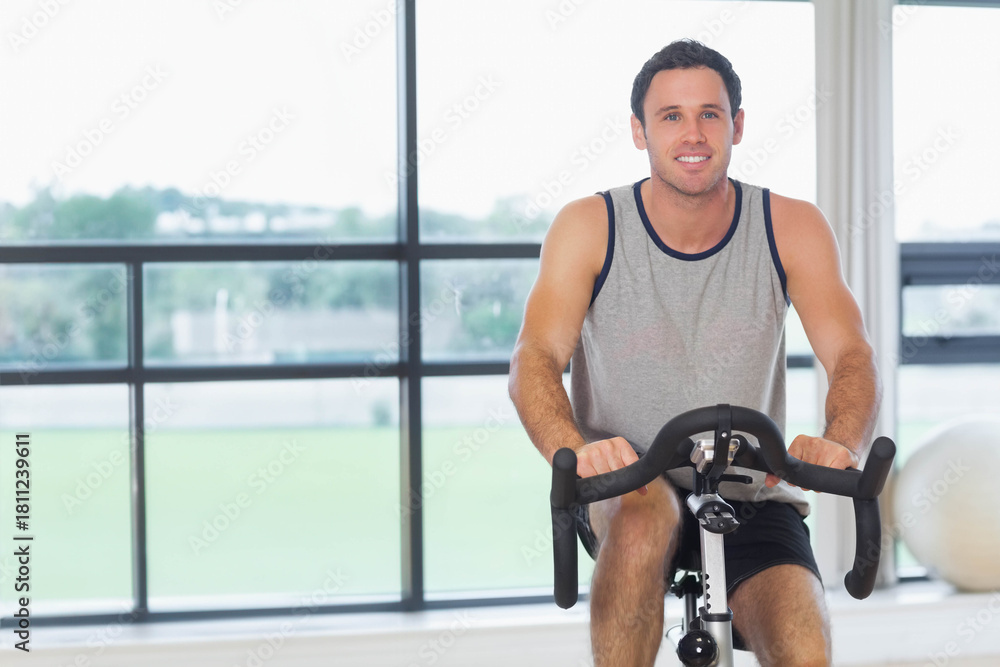 Fototapeta premium Mid-adult man cycling on stationary bike near windows at gym, wearing grey top and black shorts