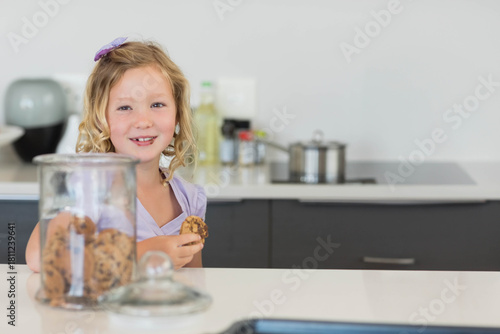Female child standing behind white counter holding choc-chip cookie, clear jar visible, purple clip