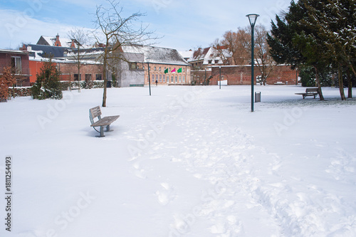 A lone park bench sits modestly in a snow-filled park, creating a calm winter moment. Snow blankets the ground near urban buildings, inviting stillness and reflection.