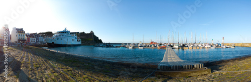 Wide harbor scene with a long pier extending into calm blue water, lined with moored boats and a distant lighthouse. Bright sun, coastal town atmosphere, and peaceful maritime activity.