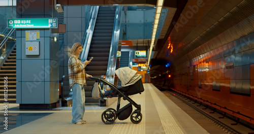 Young mother multitasking, checking smartphone while waiting with baby stroller at subway station platform, showcasing modern urban parenting lifestyle