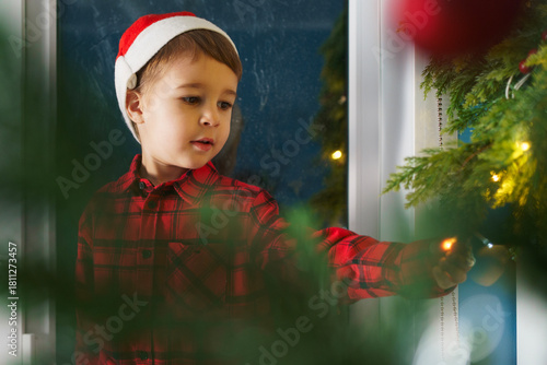 A little boy in a Santa Claus hat decorates a Christmas tree with balls and toys. 
A child in a red shirt is preparing home for the New Year celebration.