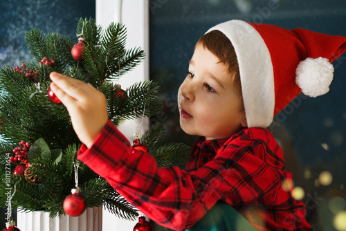 A little boy in a Santa Claus hat decorates a Christmas tree with balls and toys. 
A child in a red shirt is preparing home for the New Year celebration.