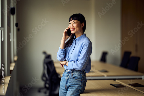 Asian Woman In Office On Phone, Smiling At Desk In Modern Workspace