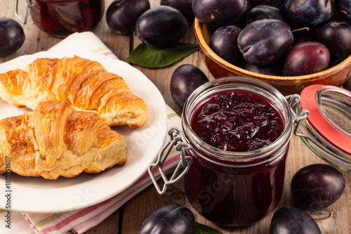 Jar of plum jam on a wooden table with fresh croissants nearby and a bowl of whole plums in the background, creating a rustic homemade food and sweet breakfast concept