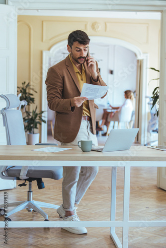 Businessman on Phone Reading Paperwork in Modern Office Desk Setup with Laptop and Coffee Workspace