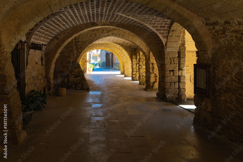 Naklejka premium Medieval stone arcade at Plaça de les Voltes in Peratallada, with vaulted arches, rustic textures, and soft sunlight entering from the plaza.