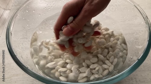 Preparing Legumes for Cooking: A close-up of a person's hand carefully washing and rinsing raw white beans in a glass bowl of clear water, the first step in making a healthy meal