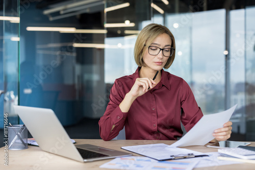 Businesswoman reviewing financial reports and data on paper, concentrating on her work at a desk with a laptop and various documents in a contemporary office environment