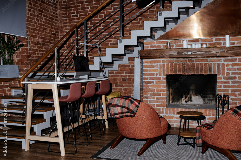 Fototapeta premium Modern business office interior showing empty workspace with computer on desk, high chairs lined up along table, brick walls, staircase, fireplace, cozy seating area, no people visible