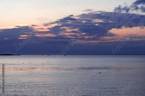 A serene coastal view at sunset, where dusky blue clouds contrast with pale pink light above the calm water. Small seabirds dot the peaceful horizon