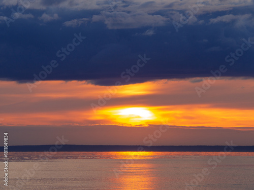 Scenic sunset over water displays vivid orange and yellow hues beneath heavy, dark clouds. The sun creates a glowing reflection on the calm surface. Dramatic cloudscape background.