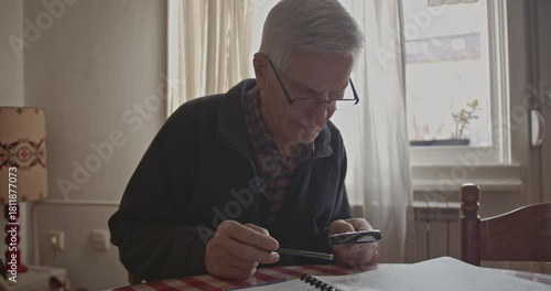 An elderly man with glasses intently reads a book or engages in a hobby at his home, using a magnifying glass to focus on the details. He appears calm and concentrated in a peaceful domestic setting.