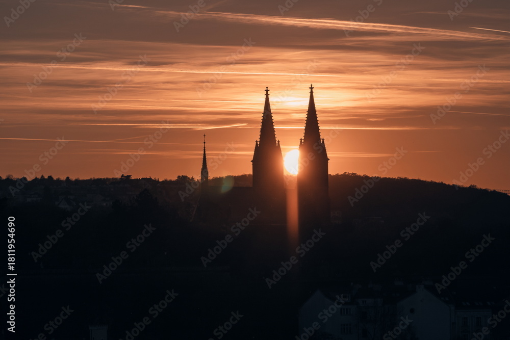 Obraz premium Dramatic sunset behind Prague’s Vyšehrad Basilica, the sun glowing perfectly between the twin spires and casting a warm orange silhouette over the historic skyline.
