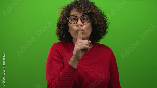 Foto Hispanic middle age woman in red sweater finger to lips for silence in studio set with bright green background; secrecy