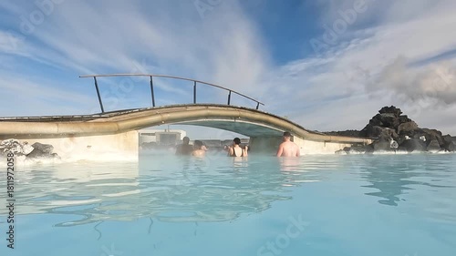 People enjoy soaking in warm, mineral-rich waters at a geothermal spa in Iceland. The sun sets, casting a beautiful glow over the steam rising from the water.
