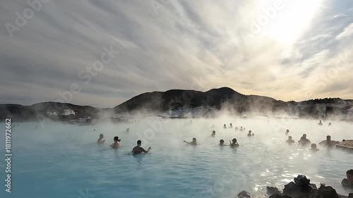 People enjoy soaking in warm, mineral-rich waters at a geothermal spa in Iceland. The sun sets, casting a beautiful glow over the steam rising from the water.
