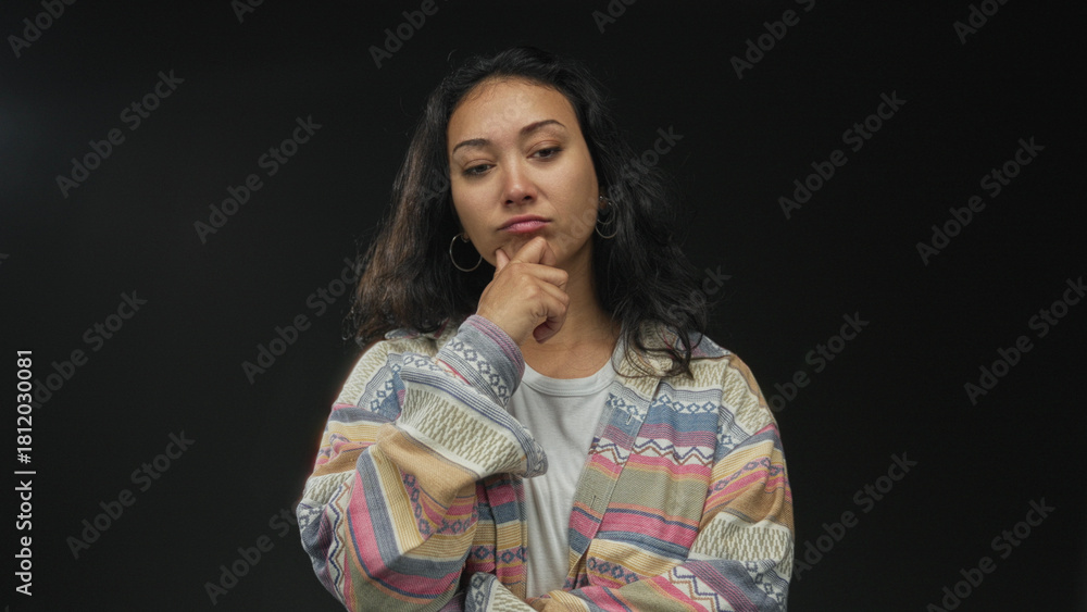 Fototapeta premium Woman with hand on chin in black studio wearing patterned cardigan and hoop earrings; thoughtful introspection.