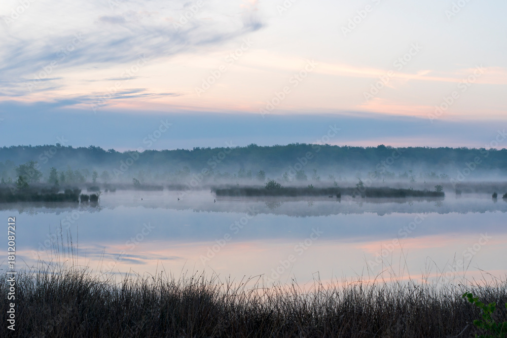 Fototapeta premium In the early morning a lake with a reflection of the sky. The sky is overcast and the air is cool and damp. The scene is quiet and peaceful, with the lake providing a sense of solitude and calm. Dwing