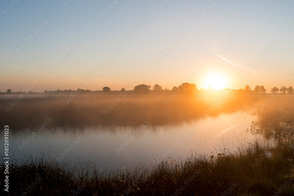 Obraz premium A sunrise over a lake in the Netherlands. The sky is orange and the sun is rising at the horizon.