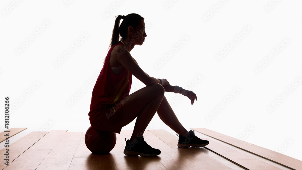 Naklejka premium Basketball player resting on ball after training in silhouette lighting. Concept of sports recovery, discipline, training focus, editorial storytelling, and mental health awareness.