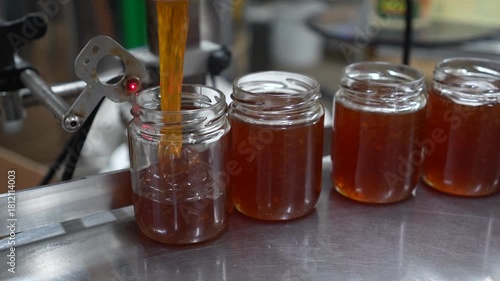Liquid honey is being poured into a glass jar on a production line, showcasing the filling process with a close-up view, emphasizing the smooth flow and texture